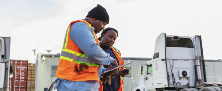 Two mature African-American workers at a freight transportation company conversing, looking at the clipboard that the man is holding. Semi-trucks and trailers with cargo containers are in the background.